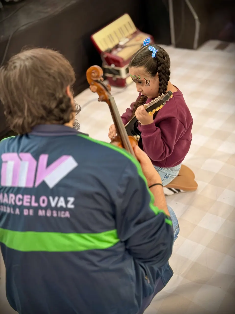 Uma menina experimenta um instrumento de cordas pequeno diante de um professor da escola, de costas, com a jaqueta da EMMV e um violino na mão.