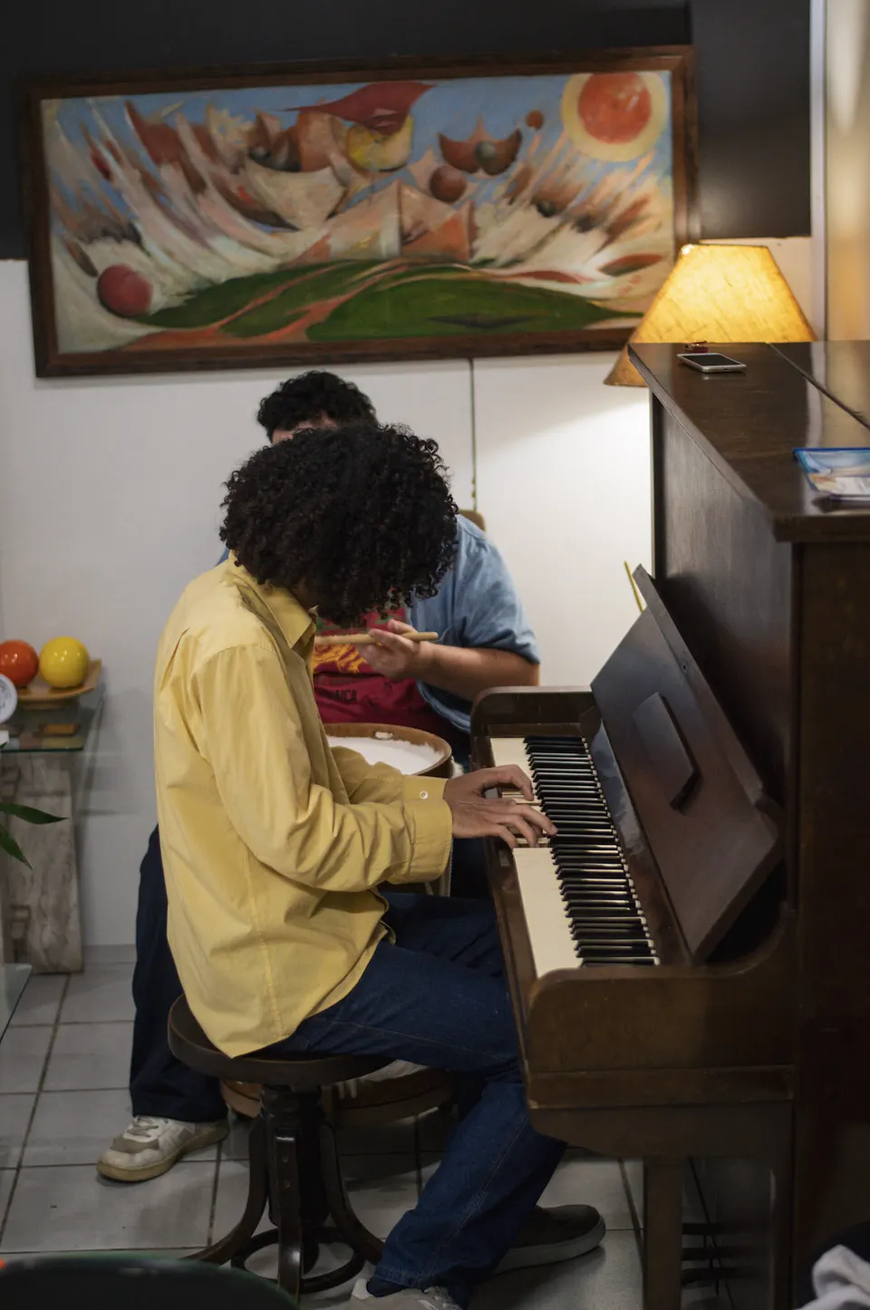 Aluno ao piano de parede da escola, sob quadros coloridos.
