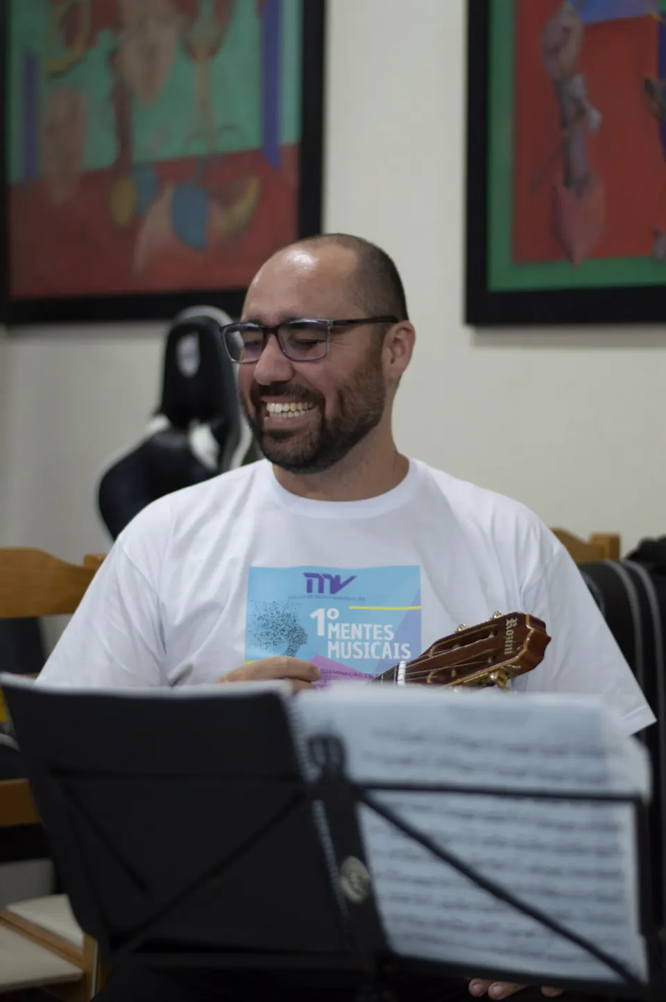 Músico com a camiseta do 1º Mentes MusiCaos sorri diante da estante de partitura.