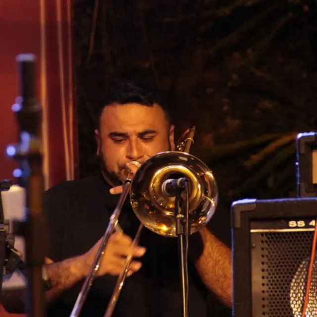 Alexandre Salum toca trombone ao microfone, num palco com cortina vermelha, de camiseta preta.