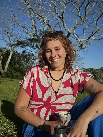 Belén Barboza Días sorri sentada na grama, ao sol, com uma cuia de chimarrão nas mãos.