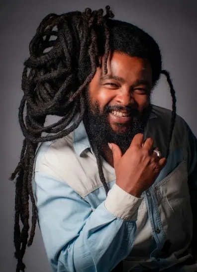 Dionísio Souza sorri com a mão no queixo, de dreadlocks longos e camisa jeans, num retrato de estúdio.