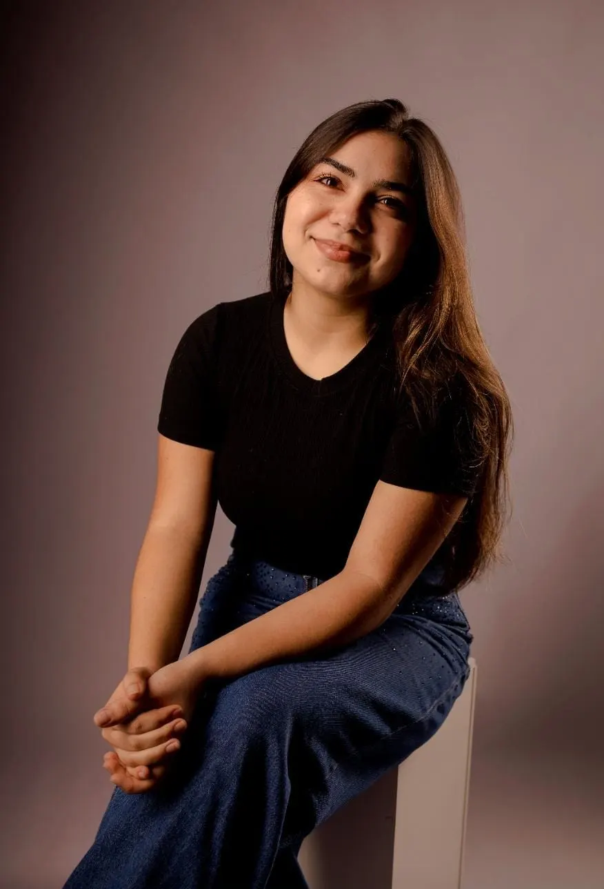 Emily Siebre sorri, sentada, de camiseta preta e calça jeans, num retrato de estúdio.