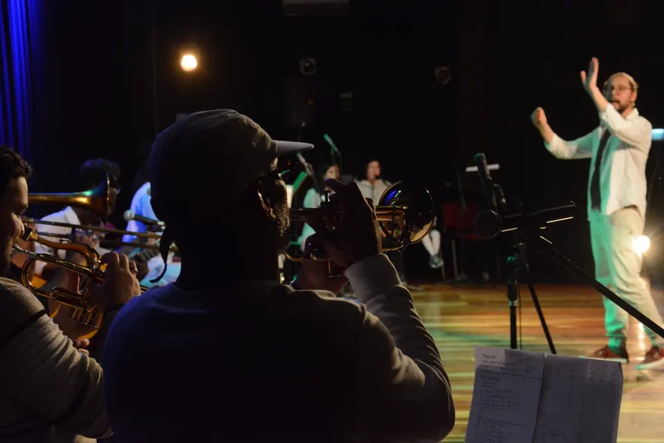 O Grupo Musical do Curso de Extensão em Música no palco: trompetes e trombone em primeiro plano e o regente, de camisa branca, ao fundo.