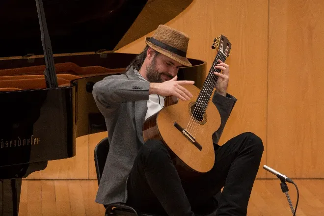 José Daniel toca violão sentado num palco, de chapéu e paletó cinza, ao lado de um piano de cauda.