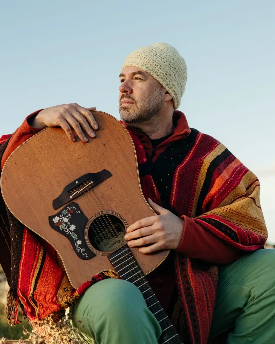 Leonardo Bulcão sentado ao ar livre, de gorro de lã e poncho listrado, abraçado a um violão, olhando para o lado contra o céu azul.