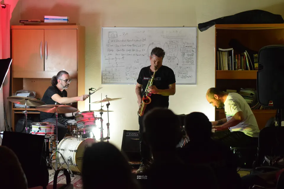 Trio de bateria, saxofone e piano toca na sala da escola, com uma lousa de harmonia ao fundo.