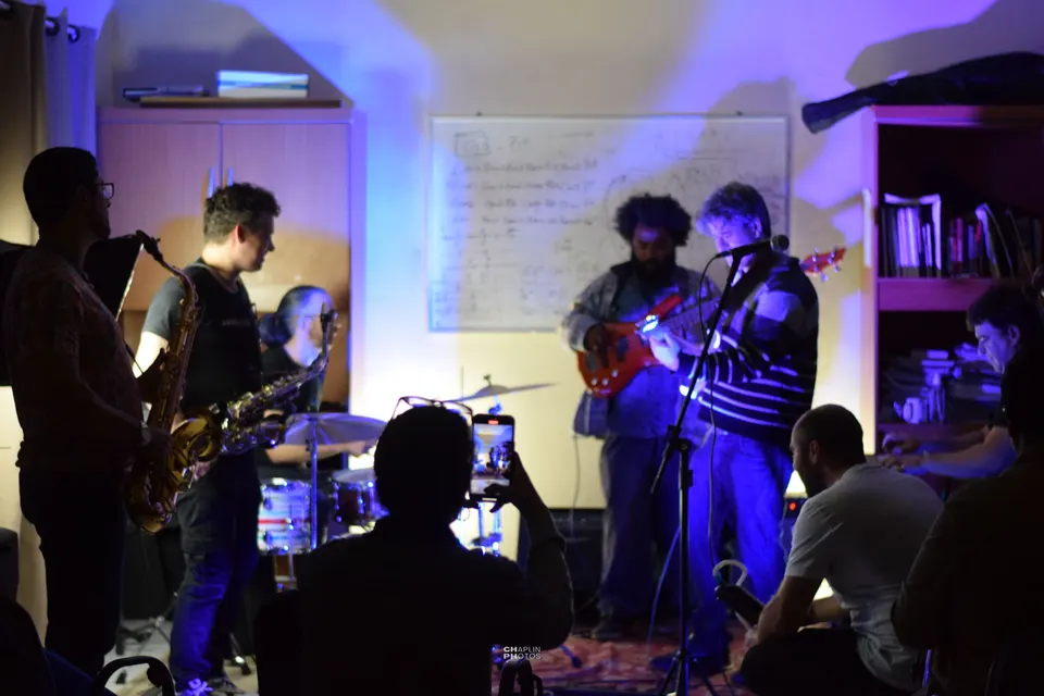 Jam session na escola: saxofones, baixo e bateria sob luz violeta, com a plateia em volta.