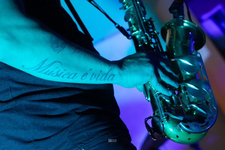 Saxofone em close sob luz ciano; no braço do músico, a tatuagem “Música é vida”.