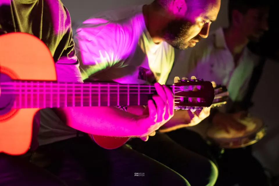 Violão tocado sob luz magenta, com outros músicos ao fundo.