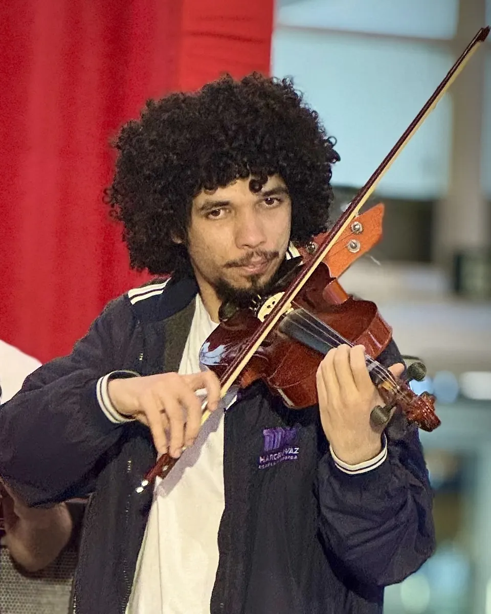 Benjamin Martins Milão toca violino num palco, de jaqueta da escola, diante de uma cortina vermelha.