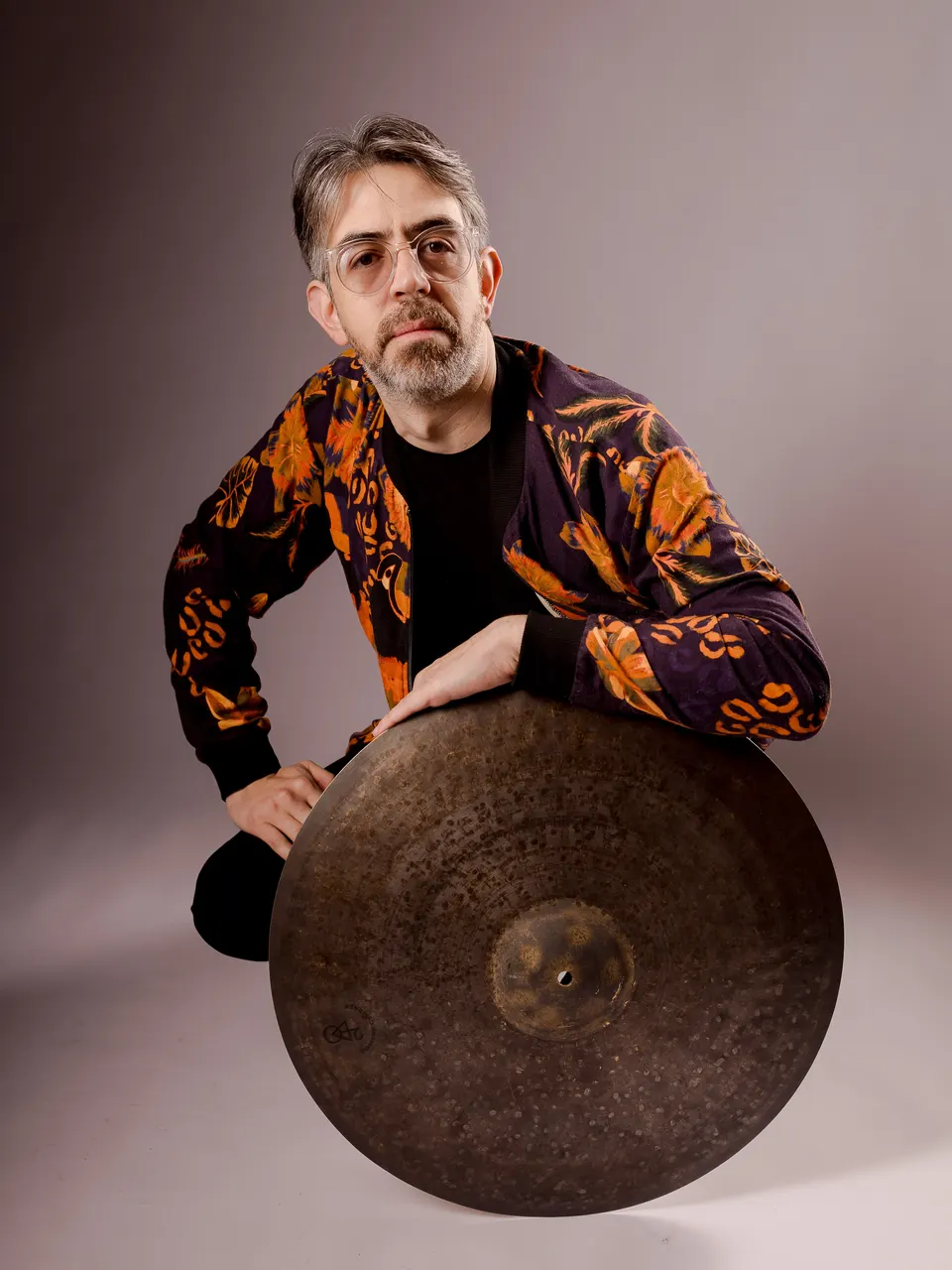 Gabriel Medeiros, de óculos de aro transparente e jaqueta estampada de flores laranja, agachado e apoiado num prato de bateria, num retrato de estúdio.