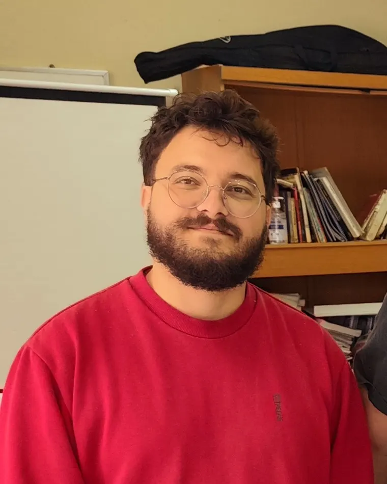 Lucas Cava, de óculos e moletom vermelho, sorri diante de uma estante de livros.