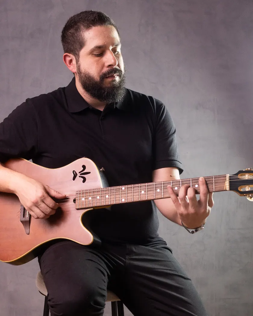 Rodrigo Arvoredo, de barba e camisa polo preta, toca violão sentado, olhando para as cordas, diante de uma parede cinza.