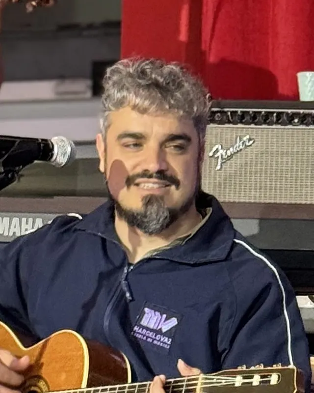 William Tavares sorri tocando violão no palco, de jaqueta da escola.
